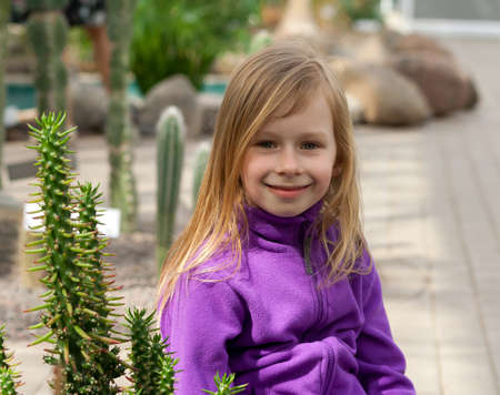 little girl of seven years in a botanical garden surrounded by a cactus, photo by waist, sitting, looking at the camera, long blond, reddish hair and gray eyes, smiling, lilac jacket background stonesの写真素材