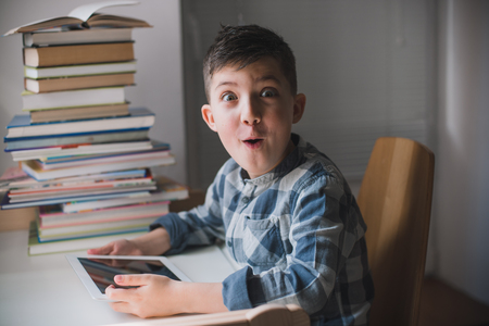 Little happy child boy holds a tablet in his hands and looks suprised sitting at a table in the background of a pile of books.の写真素材