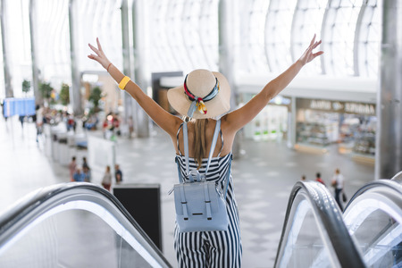Beautiful young woman at the airport descends on an escalator in a straw hat and with a backpack.の写真素材
