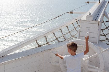 The kid in a white T-shirt holds the steering wheel of a large sailing white wooden old ship like a captain on the background of the sea.の写真素材