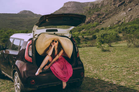 Young beautiful girl in a dress and a wide-brimmed hat near a hatchback car on a background of a mountain landscape.の写真素材