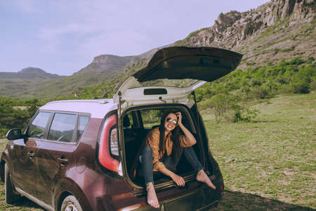 Young beautiful girl in a car trunk of a hatchback on a background of a picturesque mountain...
