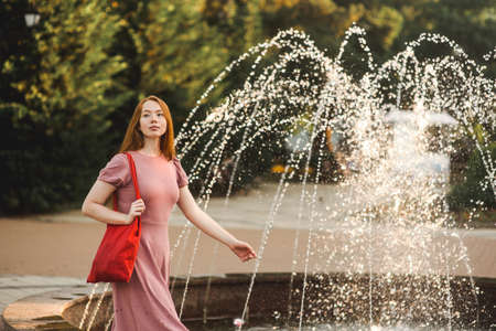 Young beautiful red-haired long-haired woman in a summer pink dress walks through the streets of a summer city.の写真素材