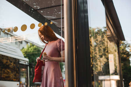 Young beautiful red-haired long-haired woman in a summer pink dress walks through the streets of a summer city.の写真素材