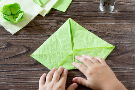 The child holds a paper square and fold it corner. The child makes crafts out of paper lily. Paper napkins on a wooden table. Children's art project, a craft for children.の写真素材