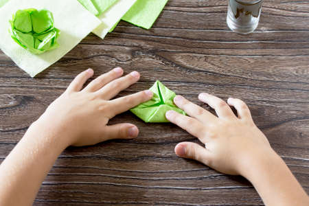 The child fold a square of paper. The child makes crafts out of paper lily. Paper napkins on a wooden table. Children's art project, a craft for children.の写真素材