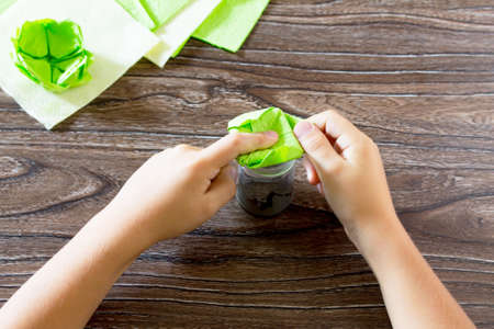 The child fold a square of paper on the glass as. The child makes crafts out of paper lily. Paper napkins on a wooden table. Children's art project, a craft for children.の写真素材