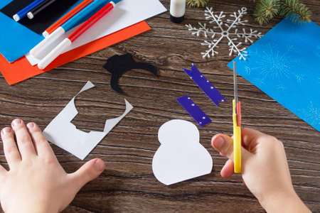 The child cut paper details snowman. The child makes a greeting card with a snowman. Glue, paper, scissors on a wooden table. Children's art project, a craft for children.の写真素材