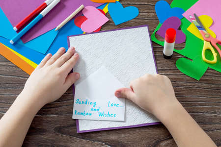 Child glues the paper items. Greeting card with paper rainbow heart. Sheets of paper, glue and scissors on a wooden table. Children's art project, a craft for children. Craft for kids.の写真素材