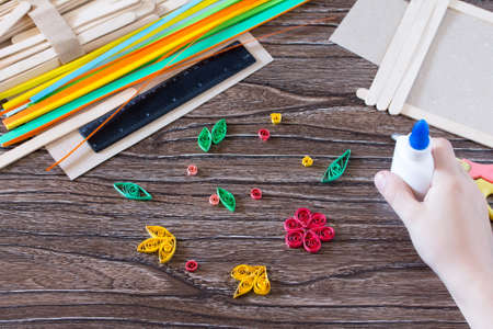 The child glues the details of the flowers in quilling technique to the gift frame of wooden sticks. Handmade. Project of children's creativity, handicrafts, crafts for children.の写真素材