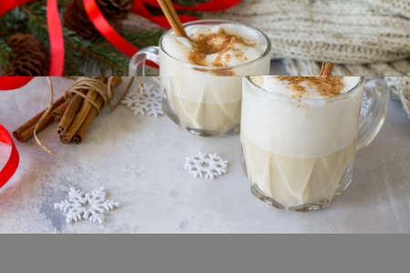 Homemade Eggnog Christmas milkshake with cinnamon, served in two cups on a gray stone or slate background, decorative ornaments, Christmas tree with copy space.の写真素材