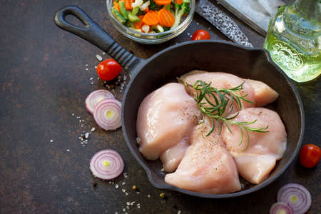 Fresh meat. Raw chicken fillet on a cast iron frying pan, spices and fresh vegetables on a kitchen table. の写真素材