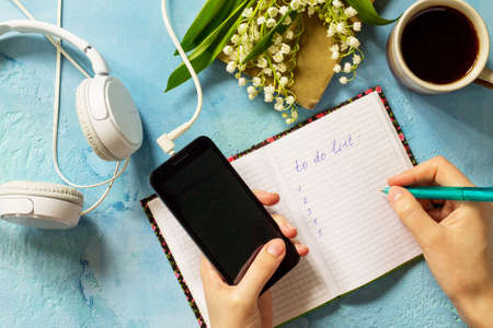 Smartphone in female hands and writing in notebook, lilies of the valley and headphones on stone table. Top view flat lay background with copy space.の写真素材