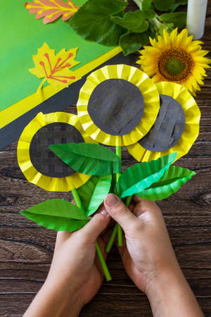 The child holds paper flower sunflower on a wooden table. Childrens art project, handmade, crafts for children.の写真素材