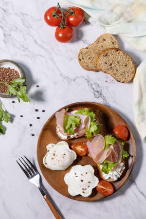 Easter breakfast or brunch. Delicious breakfast or snack - poached egg and cream cheese toast whole grain rye bread, prosciutto, arugula on a marble tabletop. Top view flat lay.の写真素材