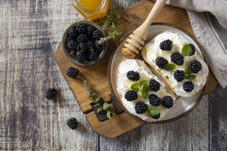 Top-down view of a rustic wooden table featuring toast with cream and fresh blackberries drizzled with honey. accompanied by a honey jar and fresh mint, creating a cozy and inviting breakfast setting.の写真素材