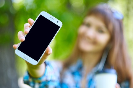 woman showing blank smart phone screen and gadget accessories on background of green foliage in...