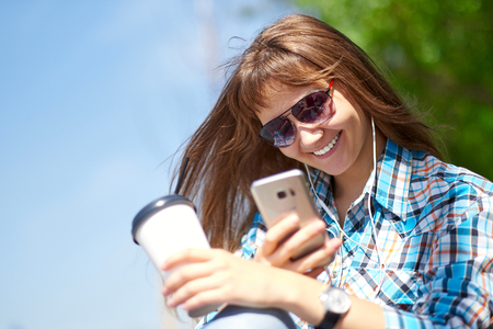 Young happy woman is using a smartphone. She is holding a paper cup of coffeeの写真素材