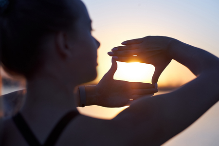 Woman silhouette framing sun with fingers at sunset. Hand shape of a camera across the skyの写真素材
