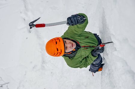 Alpinist man with ice tools axe in orange helmet climbing a large wall of ice. Outdoor Sports Portraitの写真素材