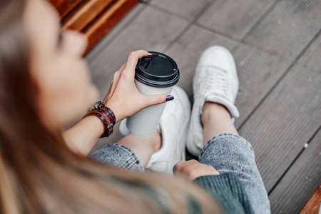 Woman in jeans sitting on a wooden bench. Holds coffee in a paper cup in hand
