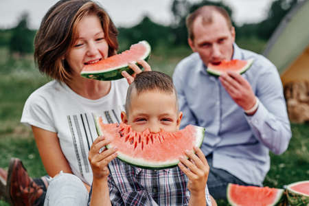 Happy family eating watermelon at picnic in meadow near the tent. Mother, father and child Enjoying Camping Holiday In Countryside.の写真素材