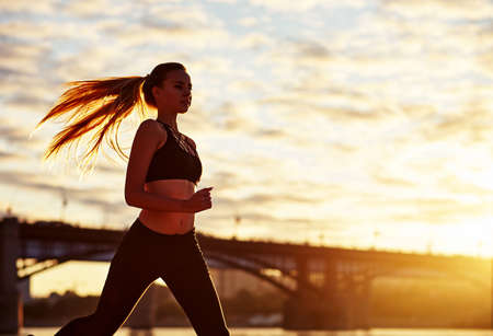 Running woman in the sand at sunrise. Morning jogging on the beach or coast of river on bridge urban city backgroundの写真素材
