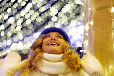 Smiling woman with garlands and holiday lights on festive Christmas or New Year fair. Lady wearing classic stylish winter knitted sweater and mittensの写真素材