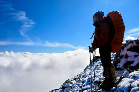 Woman climber with backpack stands on top of mountain above clouds on blue sky backgroundの写真素材