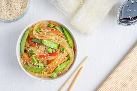 Top view of Korean Japchae savory spicy salad made of stir-fried cellophane noodles, green peas and beans with sesame seeds served in bowl with chopsticks, soy sauce and ingredientの写真素材