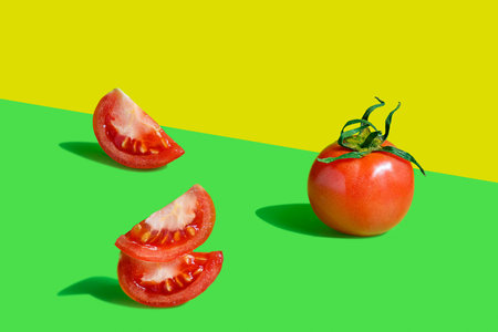 One whole red ripe juicy tomato and slices laying on bright colorful yellow and green background with geometric shadows used in healthy vegetarian or vegan dietingの写真素材