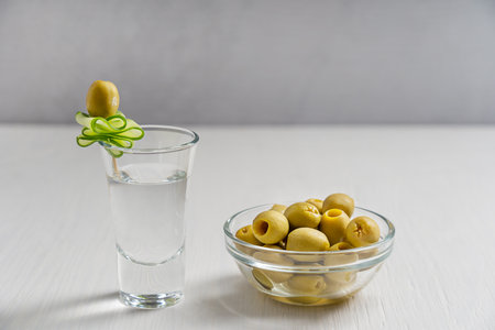 Vodka or gin clear distilled alcoholic beverage or short drink garnished with slice of raw green cucumber served in shot glass with bowl of pickled or marinated olives as snackの写真素材