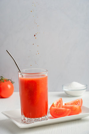 Refreshing salty tomato juice made of ripe red tomatoes served in tall drinking glass with metal straw on plate with vegetable slices on white wooden table peppering with red chiliの写真素材