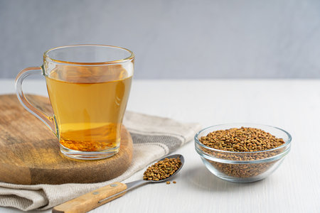Helba golden tea drink made of fenugreek annual plant served in glass cup with bowl of seeds and spoon on white wooden table with textile used in medicine of Egyptの写真素材