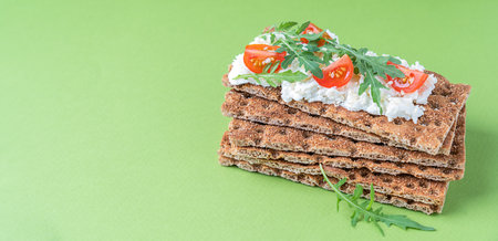 Pile of crispbread - a flat and dry type of cracker containing whole wheat flour with ricotta, cherry tomato slices and rocket leaf or arugula topping against green backgroundの写真素材