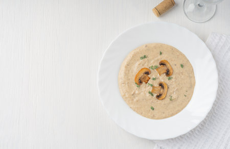 Top view of creamy mushroom soup puree made of blended champignons, potatoes and dairy cream decorated with fried slices and thyme served with glass of wine on plate on white tableの写真素材