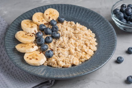 Oatmeal porridge made of boiled oats decorated with fresh blueberries and sliced banana served on blue gray plate on textile on concrete background for breakfastの写真素材