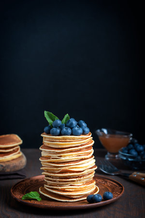 Pile of homemade sweet pancakes decorated with ripe fresh blueberries and green mint leaves topping served on plate on dark brown wooden background for breakfastの写真素材
