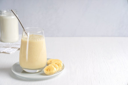 Smoothie or milkshake made of ripe sweet banana fruit blended and mixed with milk served in drinking glass with zero waste metal straw on plate on white wooden tableの写真素材