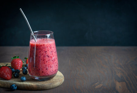 Refreshing organic smoothie made of juicy strawberries and blueberries served in drinking glass with metal reusable straw on dark brown wooden table against black wallの写真素材