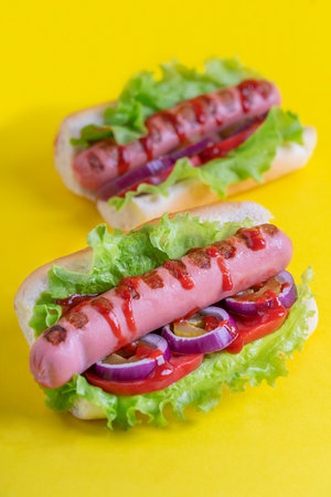 Two homemade juicy hot dogs or hotdogs made of grilled sausage with tomato, onion rings and lettuce leaves garnished with ketchup served in sliced bun on bright yellow backgroundの写真素材