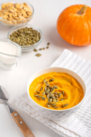 Homemade vegetarian seasonal pumpkin soup made of pureed vegetables is common Thanksgiving dish decorated with seeds served in bowl on white wooden table with spoon and napkinの写真素材