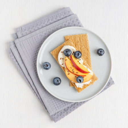 Top view of homemade sweet sandwich made with crispbread decorated with cream cheese, sliced nectarine fruit, blueberries and flax seeds served on plate on textile napkin on tableの写真素材