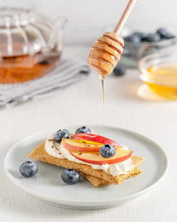 Sweet sandwich made with crispbread decorated with cream cheese, sliced nectarine fruit, blueberries and flax seeds served on plate with honey pouring from wooden dipper on tableの写真素材