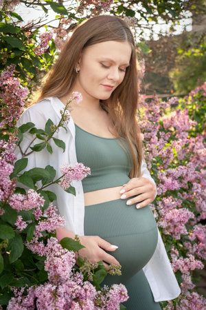 Young adult beautiful pregnant woman with blonde hair standing in front of blossoming with purple...
