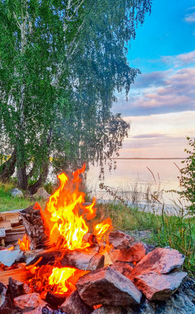Landscape of shore of a lake with idyllic place for vacation travel camping with bright glowing and burning peaceful campfire flame, green grass, birch trees, calm water and skyの写真素材