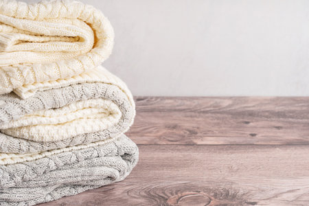 Heap or stack of folded soft cozy woven casual sweaters or cardigans of white and gray colors knitted from wool or cashmere yarn on brown wooden table clean, fresh and dryの写真素材