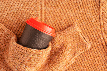 Top view of brown orange woven cozy warm textile sweater knitted from merino ot wool yarn with stripes and arm holding paper cup with hot tea or coffee drink wearing at cold winterの写真素材