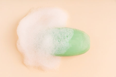 Top view of single scented antibacterial natural soap bar of green color wet and covered with white soft foam on beige background used for washing and cleaning skin at bathroomの写真素材