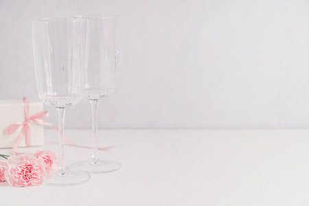 Empty transparent wine glasses served on white wooden table with copy space with pink carnation flowers and gift box with ribbon prepared for celebration of romantic valentinesの写真素材